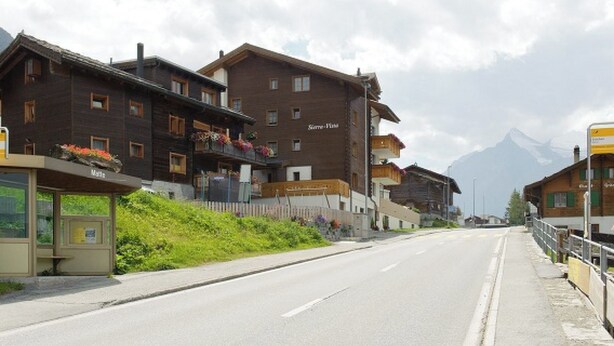 Straßenansicht mit traditionellen Holzhäusern, blauem Himmel und einem Berg im Hintergrund.