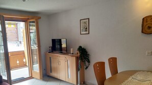 Ein Zimmer mit einem Holzschrank, einem Fernseher, einem Esstisch mit Stühlen und einer Tür, die zu einem Außenbereich führt.