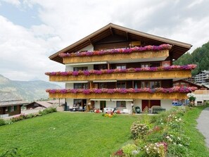 A multi-story hotel with balconies adorned with flowers, a well-maintained lawn, and mountainous scenery in the background. - Nr. 20 Sierra Vista, (3925 Grächen), 5001 by Interhome (Grächen)
