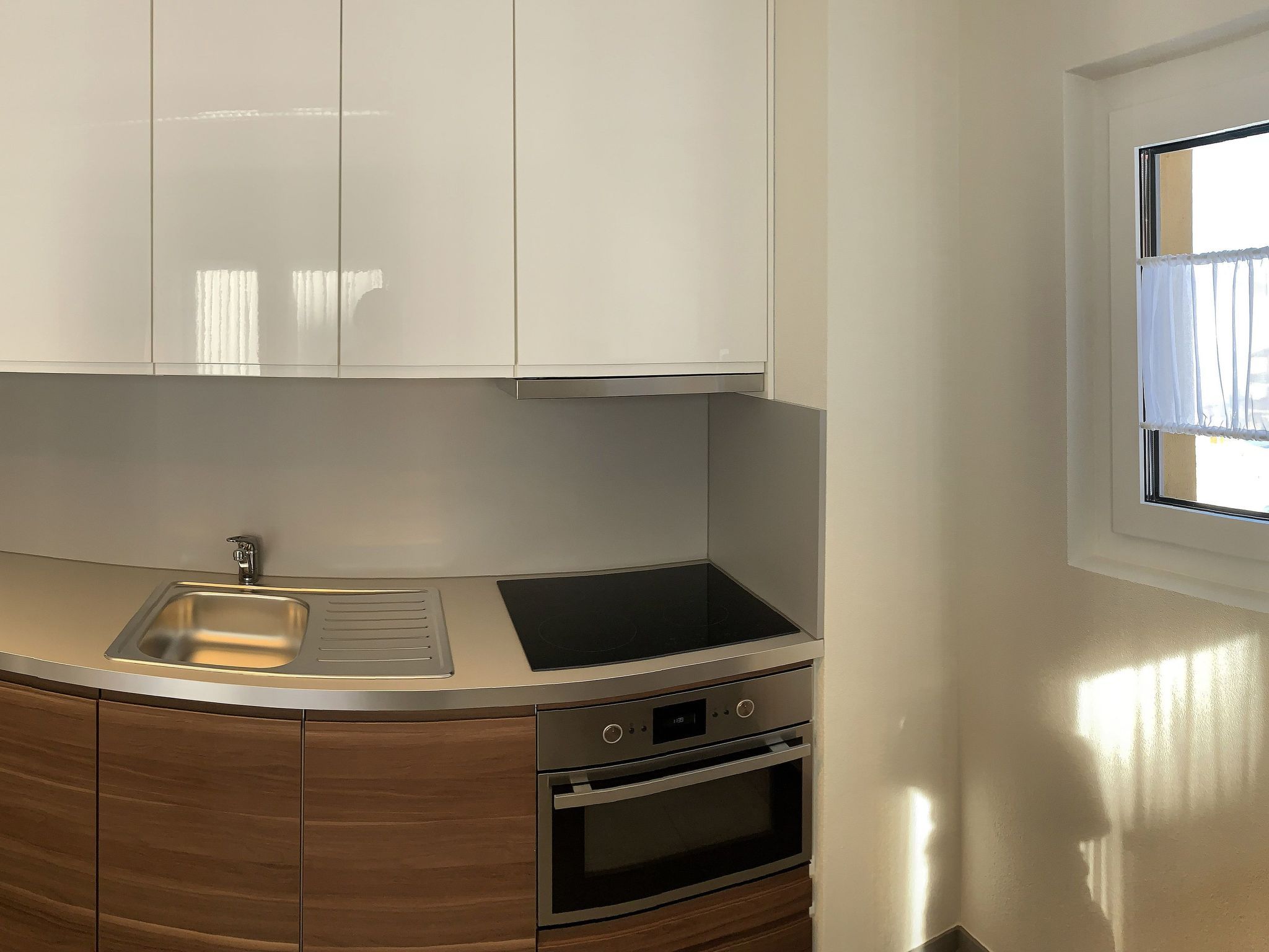 A kitchen with white cabinets, a stainless steel sink, a built-in oven, and a window with white curtains.