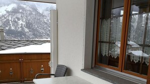 Un balcón con una mesa y dos sillas, una cerca de madera y una ventana con cortinas de encaje.