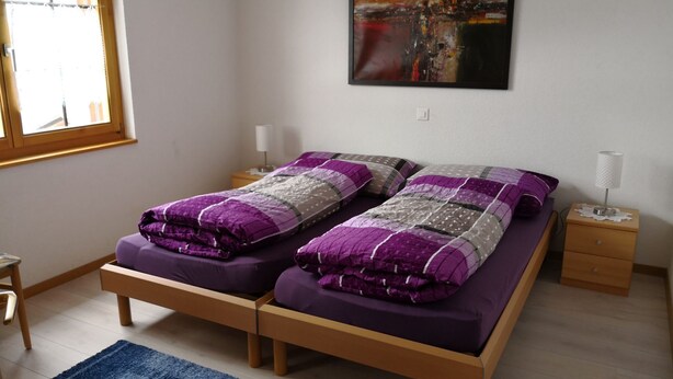 Una cama doble con colcha morada y blanca, una mesita de noche con lámpara y una ventana con persianas.