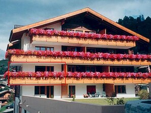 Ein mehrstöckiges Gebäude mit Balkonen, die mit roten Blumen geschmückt sind, ein flaches Dach und Blick auf eine gebirgige Landschaft.