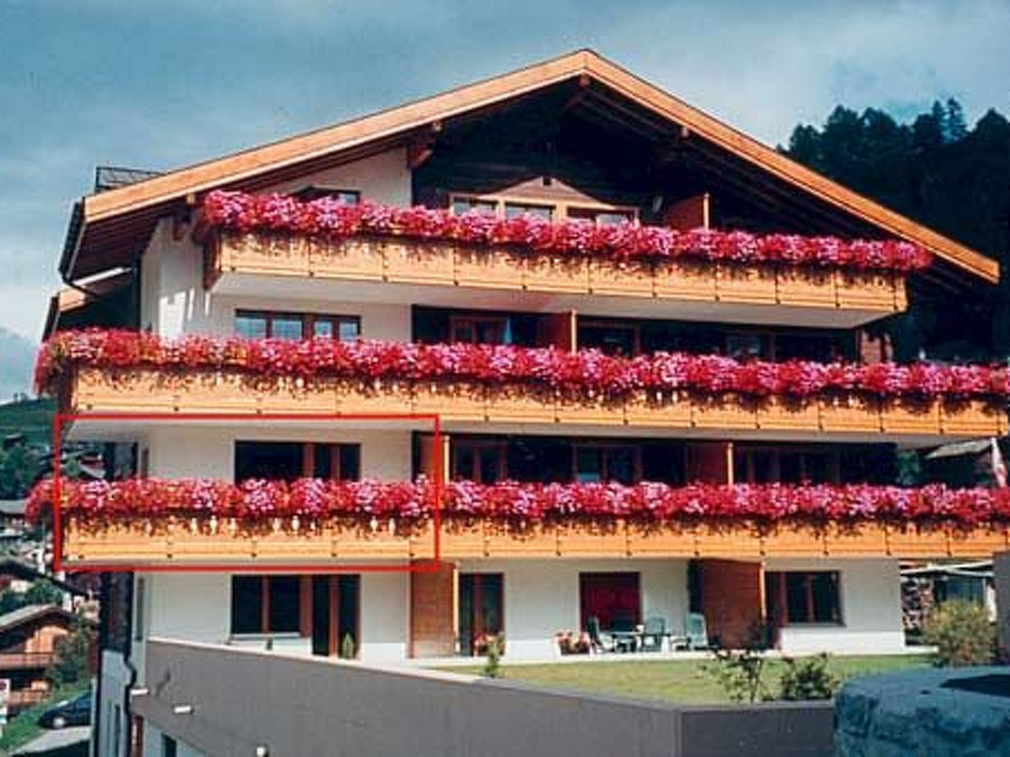 A multi-story building with balconies adorned with red flowers, a flat roof, and a clear sky.