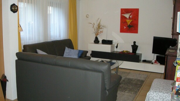 Un salón con un sofá gris, una mesa de centro, un mueble de televisión con un televisor de pantalla plana, un jarrón con plantas secas y un cuadro abstracto rojo en la pared.