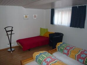 Eine kleine, einfache Unterkunft mit einem Bett, einem Sessel, einem Nachttisch und einem Fenster mit Vorhängen.