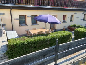 Eine Terrasse mit Tisch und Stühlen, einem violetten Sonnenschirm und einer Hecke.