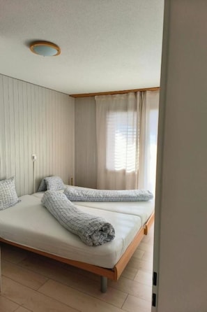 A room with two beds, a wooden headboard, a window with curtains, and a ceiling light fixture. - Ferienwohnunung Nr.22,Haus Azurit, (Grächen), 1201 by Interhome (Grächen)