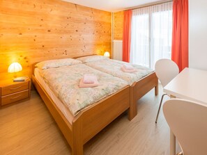 A double bed with a floral bedspread, two bedside tables with lamps, a white desk, and a chair. - Ferienwohnungen Miranda, (Grächen), by Interhome (Grächen)