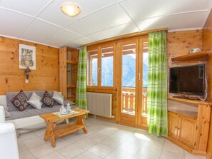 Ein gemütliches Wohnzimmer mit Holzvertäfelung, einer Couch, einem Couchtisch, einem Fernseher auf einem Holzgestell und Blick ins Freie durch eine Glastür.