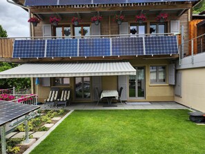 A modern two-story building with solar panels on the roof, a covered patio with outdoor furniture, and a well-maintained lawn. - Steinbock, (Grächen), 2024 by Interhome (Grächen)