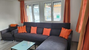 Moderni olohuone, jossa on tummanharmaa kulmasohva, oranssit koristetyynyt, lasinen sohvapöytä ja suuret ikkunat, joissa on oranssit verhot.