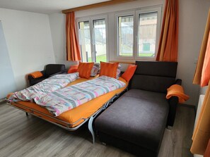 A single-bed room with a pull-out sofa, orange bedding, and a window with curtains. - Steinbock, (Grächen), 2024 by Interhome (Grächen)