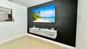 A modern living room with a large TV mounted on a black wall, a white entertainment unit below, and a kitchen area in the background. - Tropical Bliss Retreat: Poolside Paradise Awaits (Pembroke Pines)
