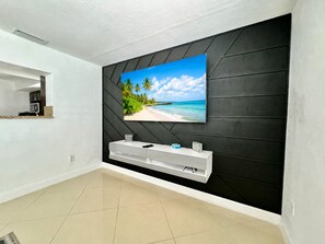 Ein modernes Wohnzimmer mit einem großen Fernseher an einer schwarzen Wand, einer weißen Unterhaltungseinheit darunter und einer Küche im Hintergrund.