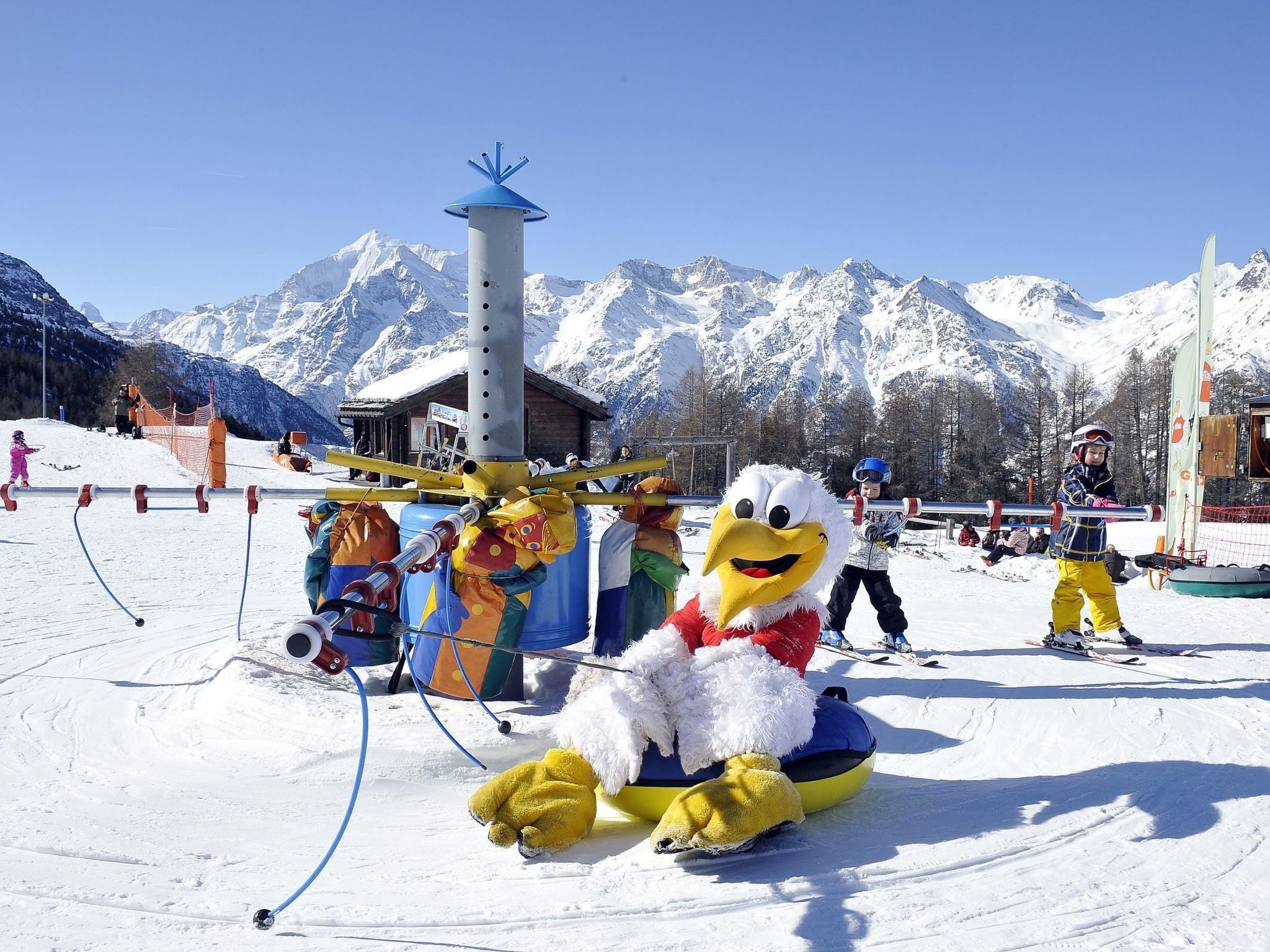 En snötäckt skidort med en maskot, en skidlift och en bergskedja i bakgrunden.