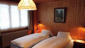 Une chambre avec deux lits, un radiateur, une fenêtre avec des rideaux, un tableau accroché au mur et un luminaire suspendu.