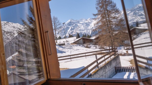 A view through a window with a wooden frame, snow-covered ground, a wooden staircase, and a mountain in the background. - Dominique, (Grächen), 1013 by Interhome (Grächen)