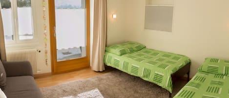 Una habitación con sofá, dos camas individuales con ropa de cama verde, una ventana con cortinas y un radiador.