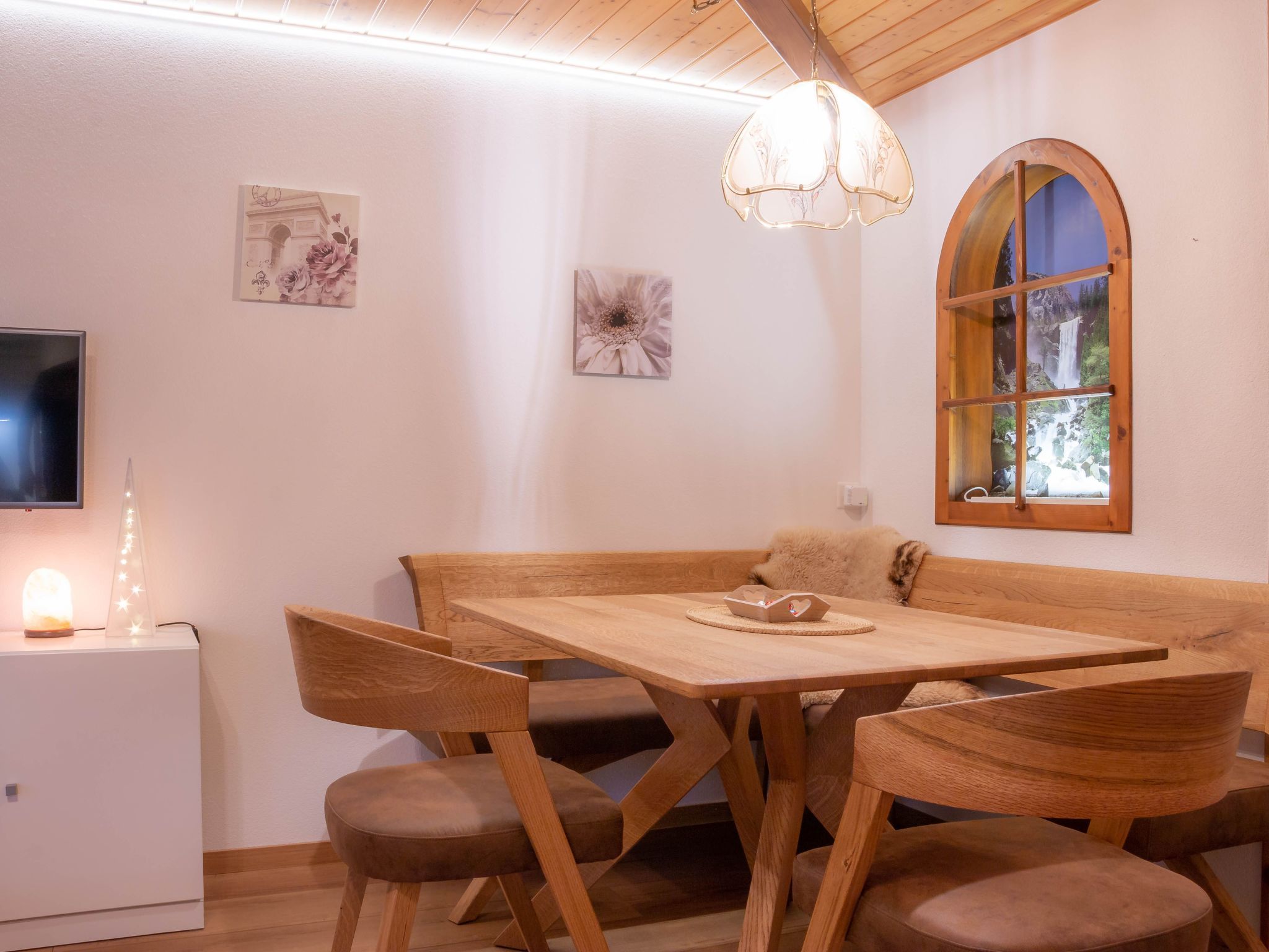 A dining area with a wooden table and chairs, a bench, a decorative light, and a window with a view of a waterfall.