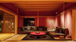 Un salon de style japonais traditionnel, avec un plafond bas en bois, un canapé en cuir marron, une table basse ronde rouge et un sol recouvert de tatamis.