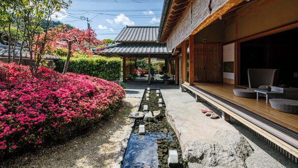 Il s’agit d’un bâtiment japonais traditionnel, avec une façade en bois, un toit recouvert de tuiles et un jardin agrémenté de fleurs roses et d’un sentier en pierre.