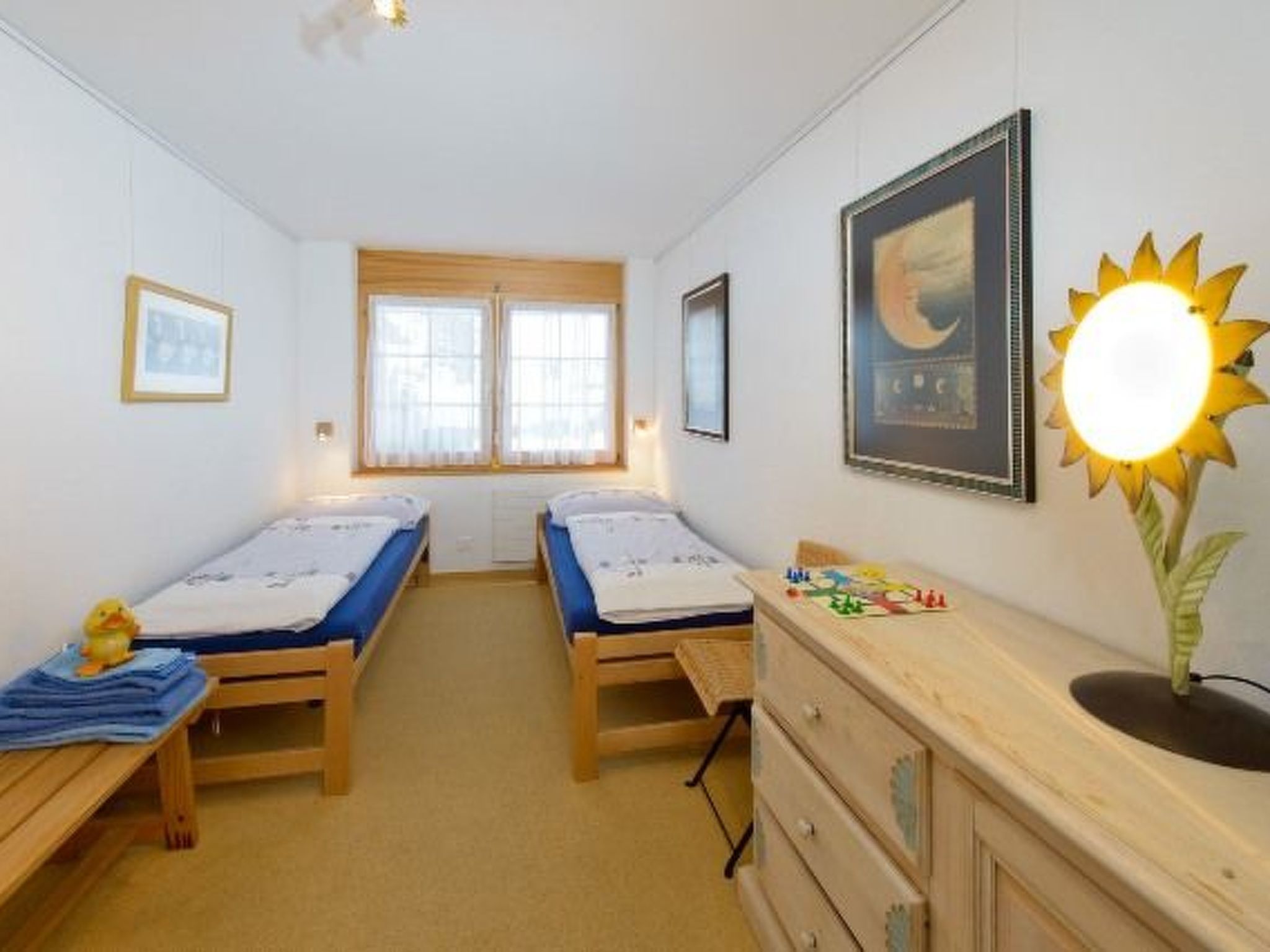 A bedroom with two single beds, a dresser, a window with blinds, and a wall-mounted lamp.