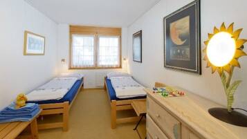 A bedroom with two single beds, a dresser, a window with blinds, and a wall-mounted lamp.