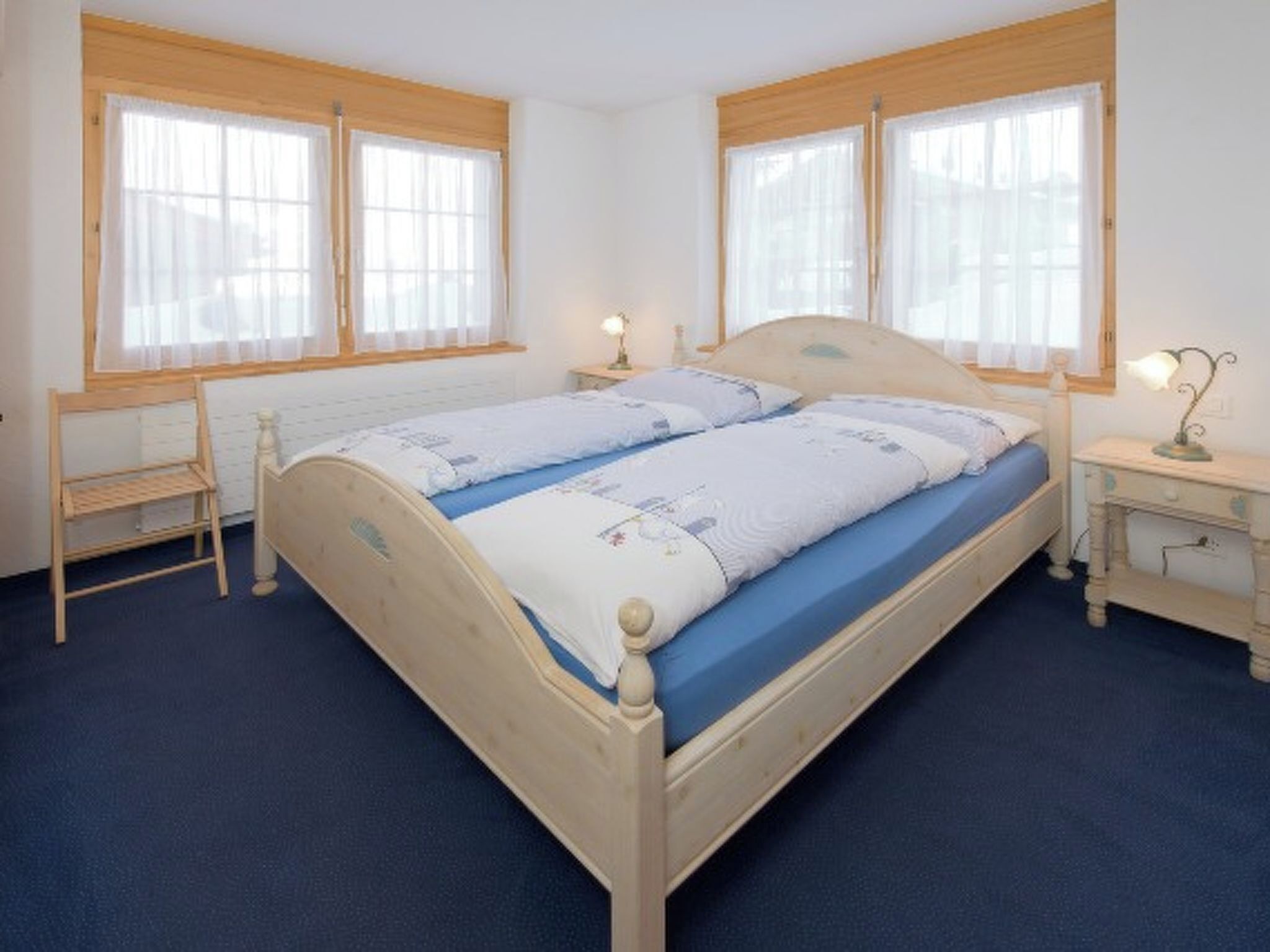 Ein Doppelbett mit blauer Matratze, zwei Nachttische mit Lampen, eine Holzbank und große Fenster mit durchsichtigen Vorhängen.