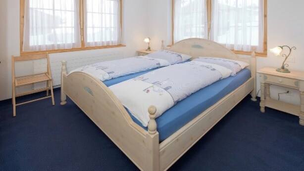 Ein Doppelbett mit blauer Matratze, zwei Nachttische mit Lampen, eine Holzbank und große Fenster mit durchsichtigen Vorhängen.