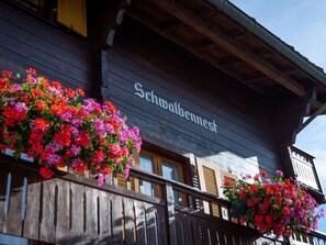 Un bâtiment dont la façade porte l’inscription « Schmalbennest », des balcons en bois ornés de jardinières fleuries et un ciel dégagé.