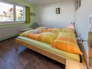 Ein Zimmer mit einem hölzernen Bett, einem Heizkörper, einem mit Vorhängen versehenen Fenster, einem Stuhl und einer Wandleuchte.