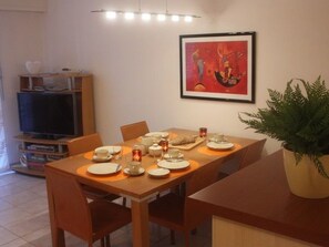 Un comedor con una mesa dispuesta para cuatro personas, un televisor sobre un mueble bajo y una obra enmarcada en la pared.