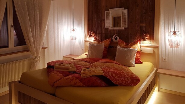 A bedroom with a bed, bedside lamps, a window with curtains, and a wooden accent wall. - Silberblick, (Grächen), by Interhome (Grächen)