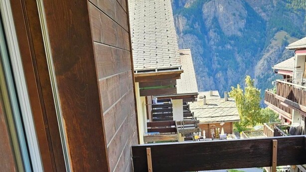 Uma varanda com mesa e banco de madeira, um pequeno recipiente de metal e vista para uma paisagem montanhosa.