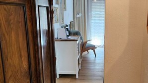Corredor que dá para um quarto com uma escrivaninha branca, uma cadeira e uma janela com cortinas.