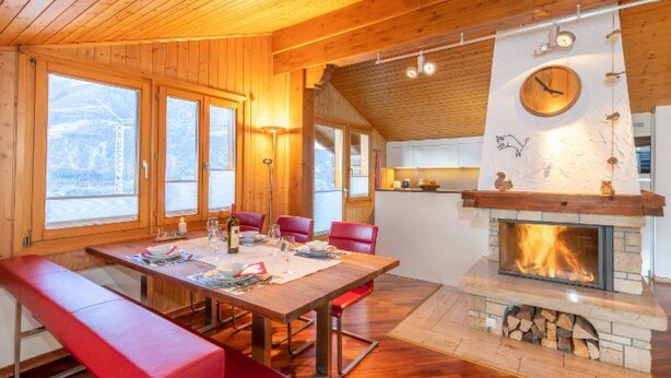 Interior acogedor de una cabaña con una mesa de comedor de madera, bancos rojos, una chimenea y ventanas amplias.