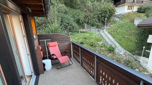 Un balcón con zona de estar, vistas a una ladera boscosa y una escalera que lleva a un edificio.