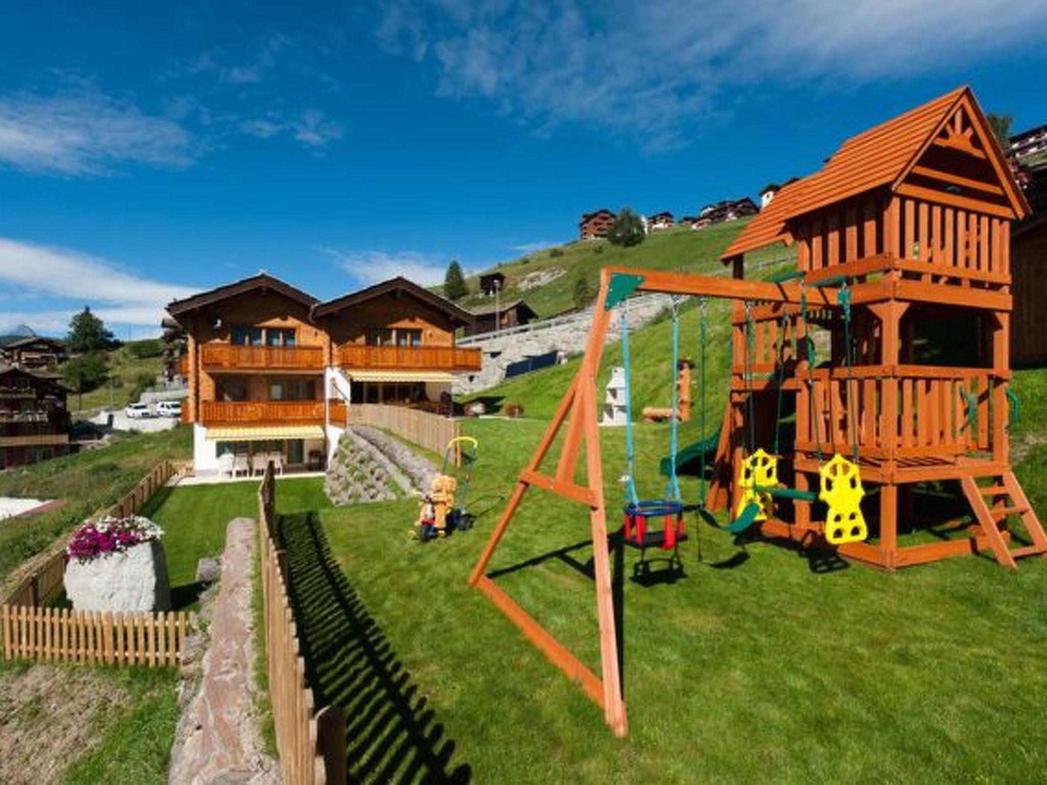 Un chalet en bois doté d’une balançoire et d’un petit toboggan, entouré d’une pelouse bien entretenue et d’un mur en pierre.