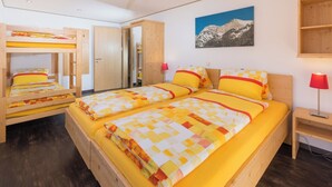Una stanza con due letti a castello, un letto singolo, mobili in legno, un quadro raffigurante montagne e una lampada rossa.