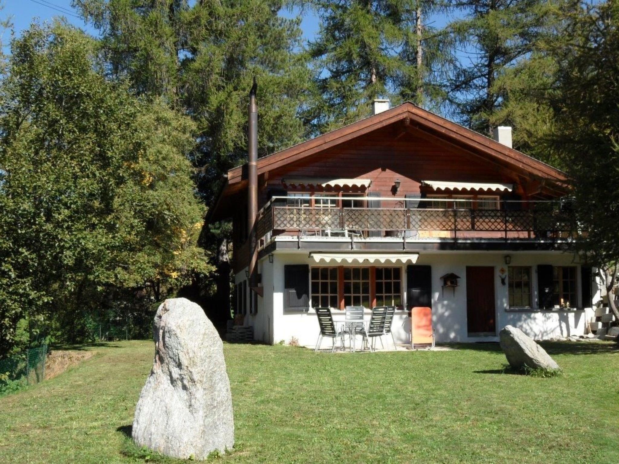 A two-story wooden house with a balcony, surrounded by trees and a grassy yard.