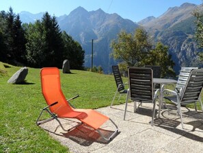 Eine Terrasse mit Gartenmöbeln, darunter ein einzelner orangefarbener Liegestuhl und ein Tisch mit vier Stühlen, vor einer Bergkulisse.