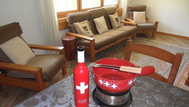 Una sala de estar con un sofá, sillas, una mesita con una botella roja y una bandera suiza, además de una alfombra estampada.