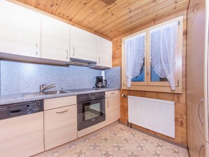 Eine Küche mit Holzschränken, einem Geschirrspüler, einem Backofen, einer Kaffeemaschine und einem Fenster mit Spitzenvorhängen.