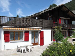 Ein zweistöckiges Gebäude mit Balkon, roten Fensterläden und einer kleinen Außen-Sitzgelegenheit.