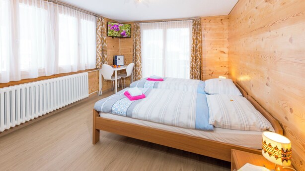 Quarto com cama de madeira, uma escrivaninha, uma cadeira, radiador e televisão na parede.