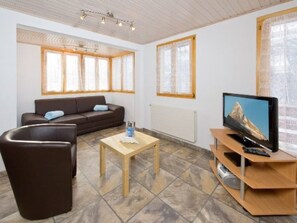 A living room with a sofa, a coffee table, a TV on a wooden stand, and large windows with curtains. - Ferienwohnungen Haus Florida, (Grächen), by Interhome (Grächen)