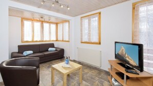 Una sala de estar moderna con sofá, una mesa de centro, un televisor sobre un mueble de madera y ventanas grandes con cortinas.