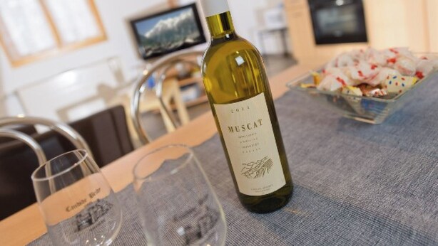 Una botella de vino Moscatel, una copa y un bol de aperitivos sobre una mesa en una habitación con cocina al fondo.