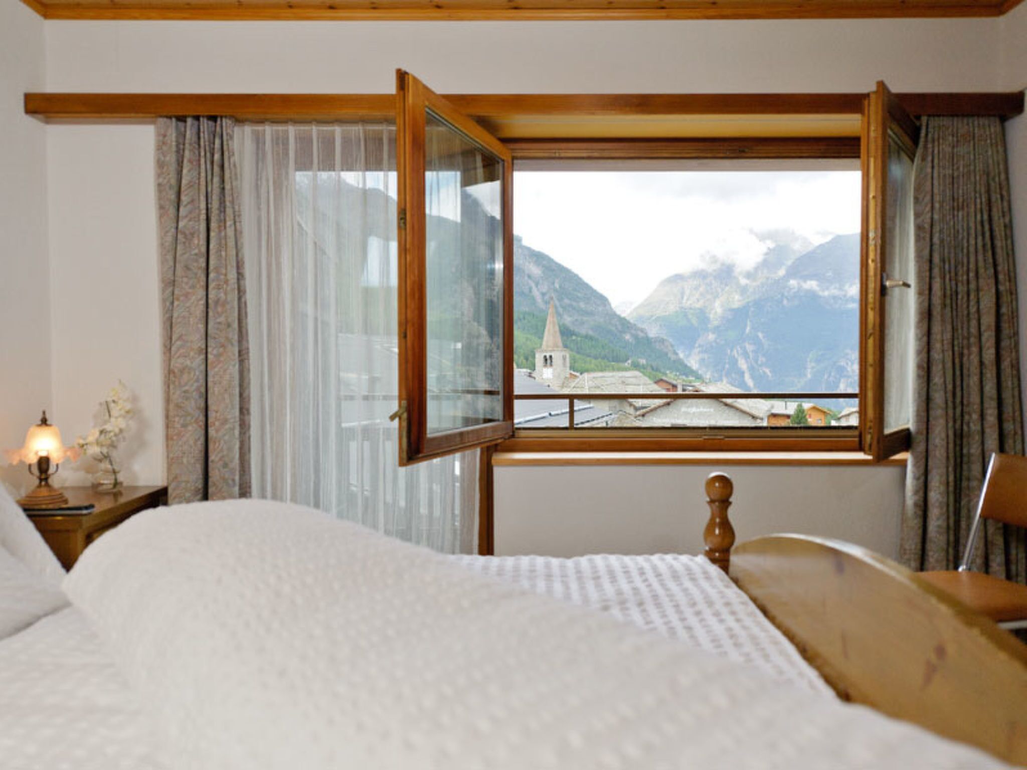A bedroom with a bed, a nightstand with a lamp, a wooden headboard, and a window with a view of mountains and a church.